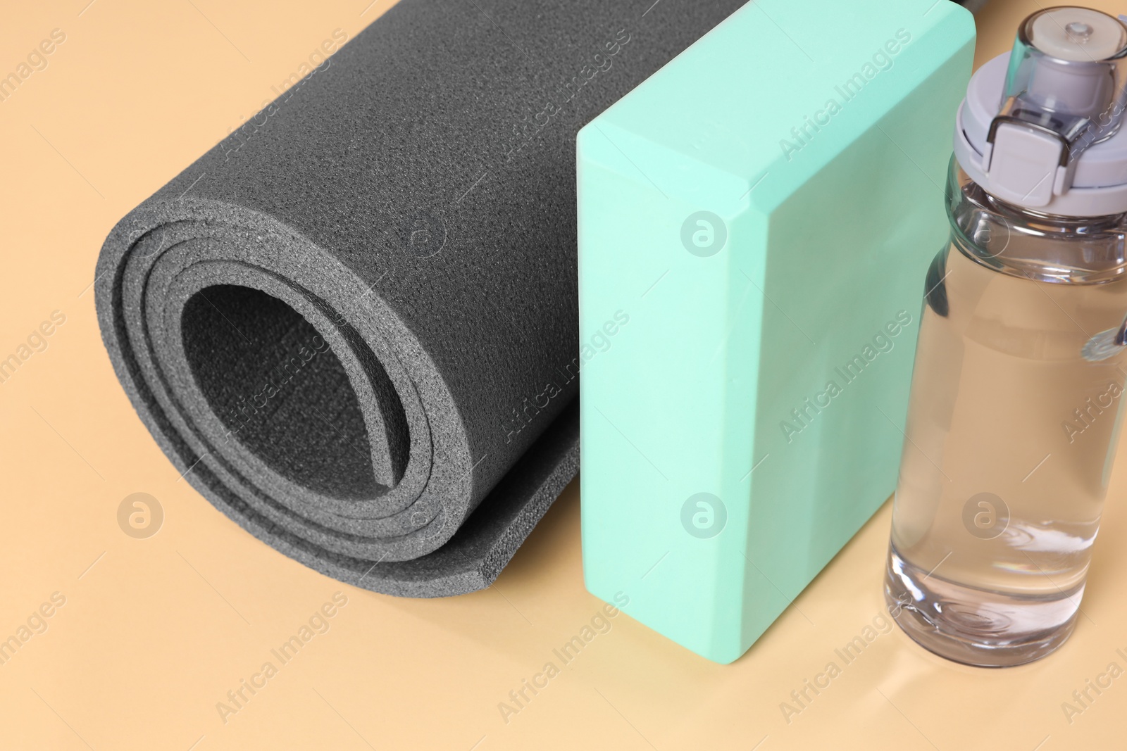 Photo of Grey exercise mat, yoga block and bottle of water on beige background, closeup