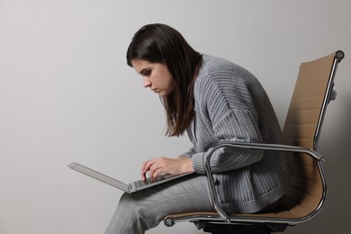 Photo of Young woman with bad posture using laptop while sitting on chair against grey background