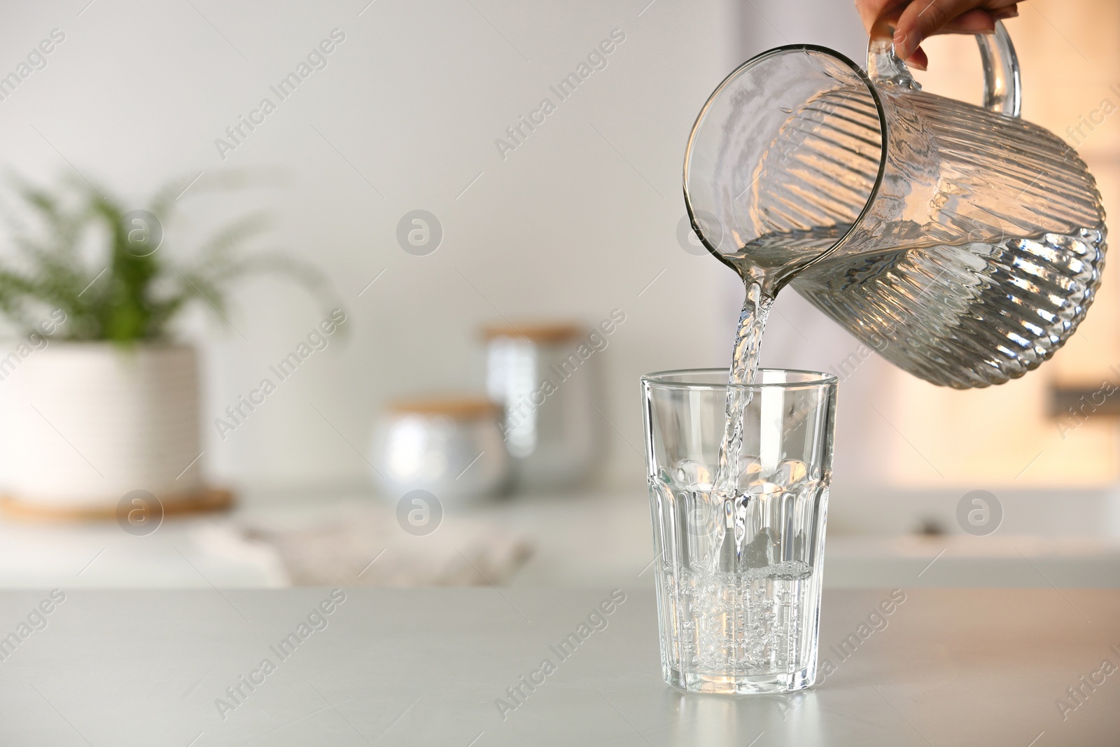 Photo of Woman pouring water from jug into glass at white table in kitchen, closeup. Space for text