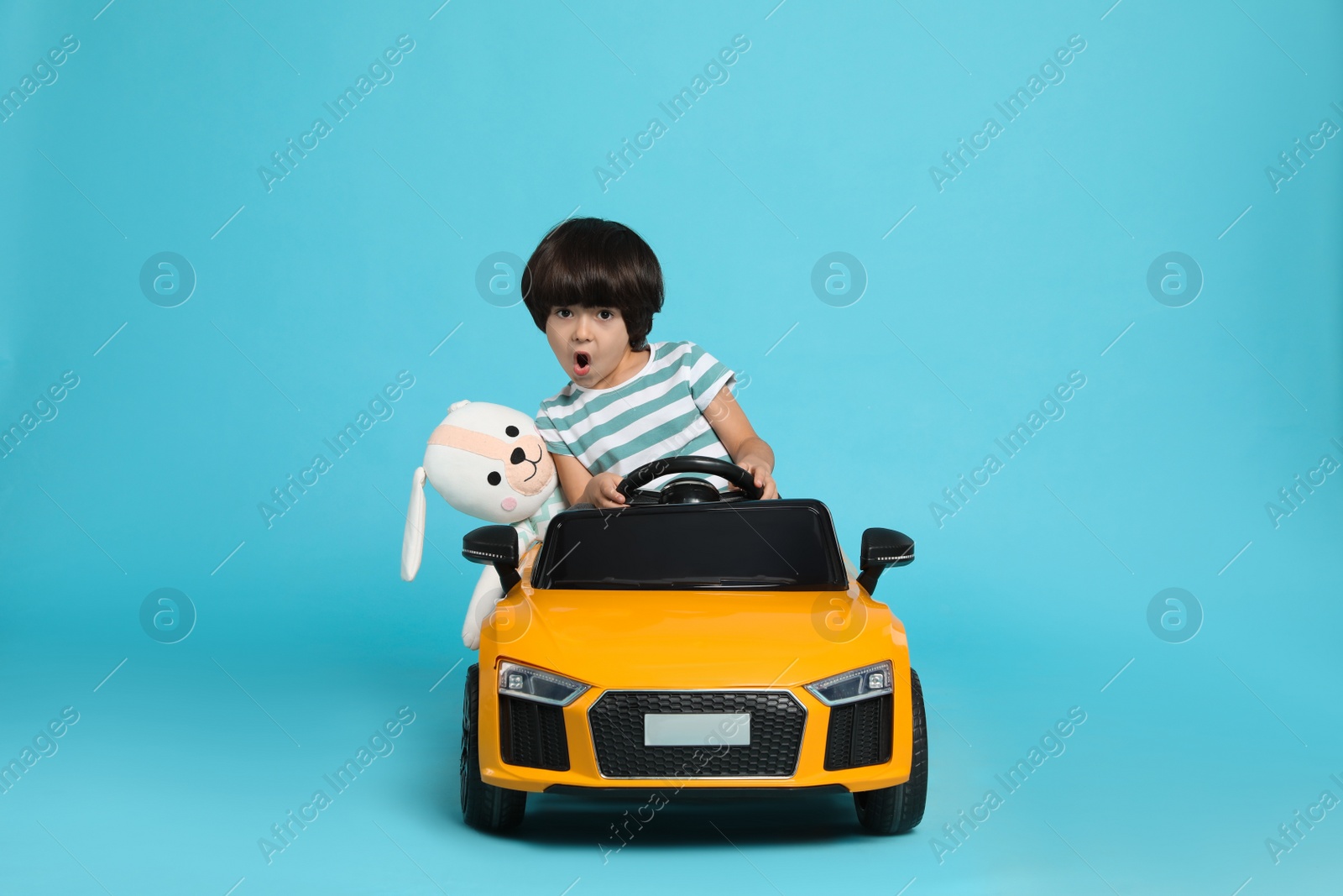 Photo of Cute little boy with toy bunny driving children's car on light blue background