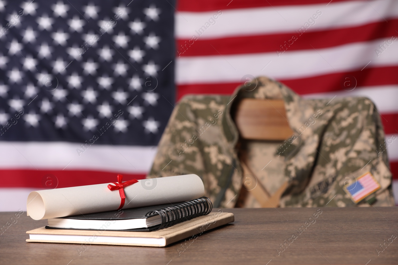 Photo of Notebooks and diploma on wooden table, chair with soldier uniform against flag of United States indoors. Military education