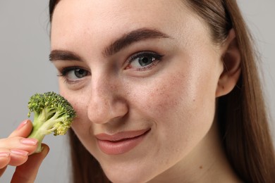Beautiful woman making fake freckles with broccoli and cosmetic product on grey background, closeup