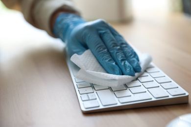 Photo of Woman in latex gloves cleaning computer keyboard with wet wipe at table, closeup
