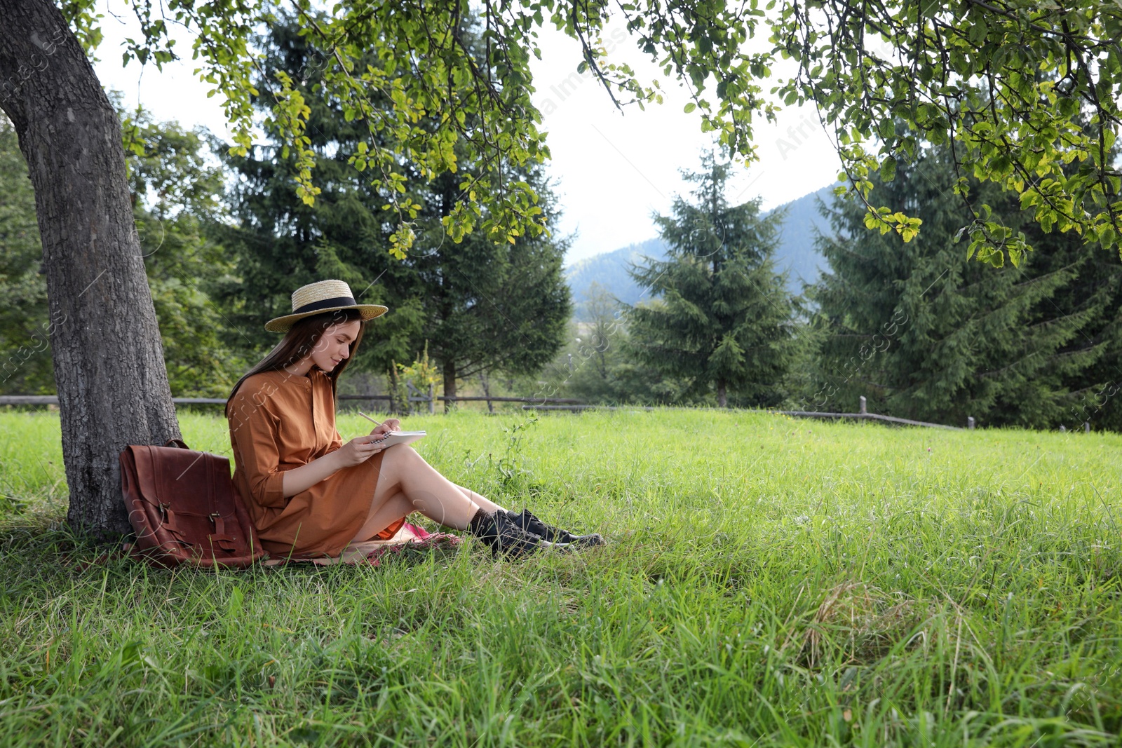 Photo of Beautiful young woman drawing with pencil in notepad near tree on green grass