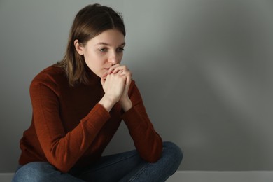 Sad young woman near grey wall indoors, space for text