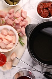 Fondue pot with oil, forks, raw meat pieces, glass of red wine and other products on white marble table, flat lay