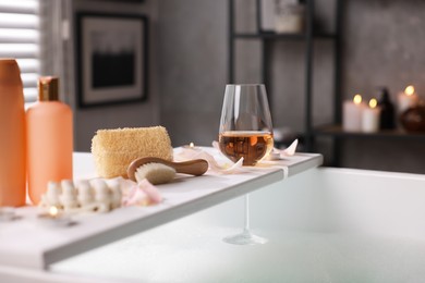Wooden tray with wine, toiletries and flower petals on bathtub in bathroom