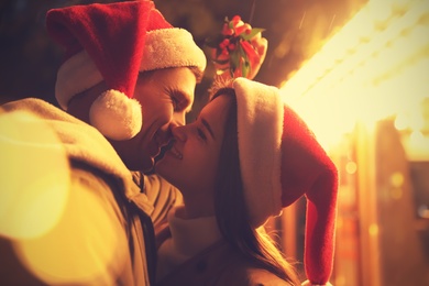 Happy couple in Santa hats kissing under mistletoe bunch outdoors, bokeh effect