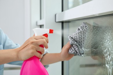 Photo of Woman cleaning window with rag and detergent indoors, closeup