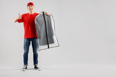 Dry-cleaning delivery. Happy courier holding garment cover with clothes and showing thumbs up on light grey background, space for text