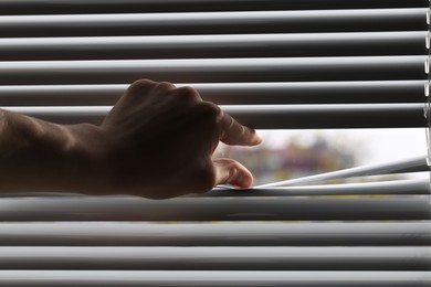 Man separating slats of white blinds indoors, closeup