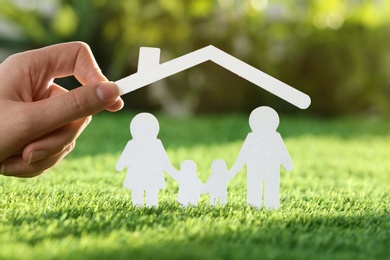 Photo of Woman holding paper roof over cutout of family on fresh grass, closeup. Life insurance concept