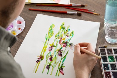 Photo of Man painting flowers with watercolor at wooden table, closeup. Creative artwork