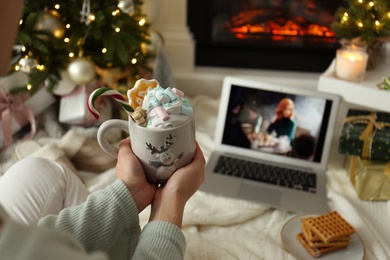 Photo of MYKOLAIV, UKRAINE - DECEMBER 23, 2020: Woman with sweet drink watching The Queen's Gambit series on laptop near fireplace at home, closeup. Cozy winter holidays atmosphere