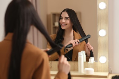 Photo of Beautiful happy woman using hair iron near mirror in room
