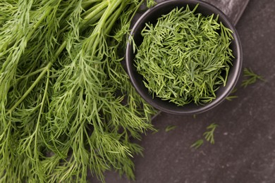 Photo of Sprigs of fresh dill and bowl with cut one on dark textured table, top view