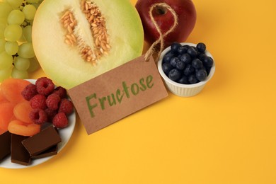 Photo of Label with word Fructose, delicious fruits, berries and pieces of chocolate on yellow background, above view