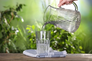 Photo of Woman pouring water from jug into glass on wooden table outdoors, closeup