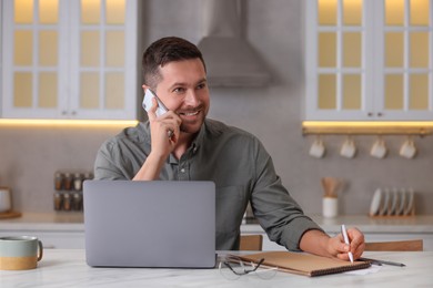 Happy man talking on phone while working with laptop at white marble table in kitchen