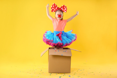 Photo of Little girl with large bow and clown nose jumping out of cardboard box on yellow background. April fool's day