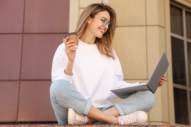Image of Young woman with paper cup of coffee working on laptop outdoors