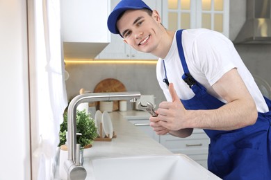 Photo of Smiling plumber repairing faucet with spanner and showing thumb up in kitchen
