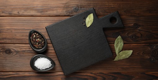 Photo of Black cutting board, salt, pepper and bay leaves on wooden table, flat lay. Space for text