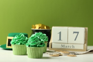 Photo of St. Patrick's day party. Tasty cupcakes with green cream, pot of gold, wooden block calendar and leprechaun hat on white table, closeup