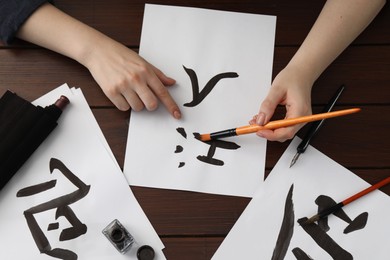 Calligraphy. Woman with brush and inkwell writing hieroglyphs on paper at wooden table, top view