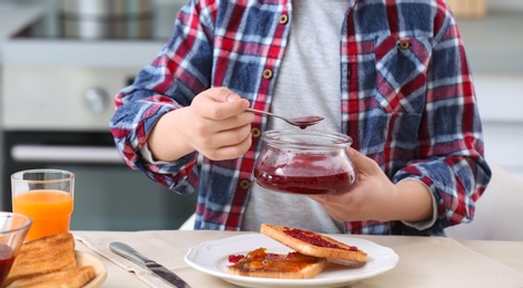 Photo of Cute little boy spreading jam onto tasty toasted bread at table in kitchen