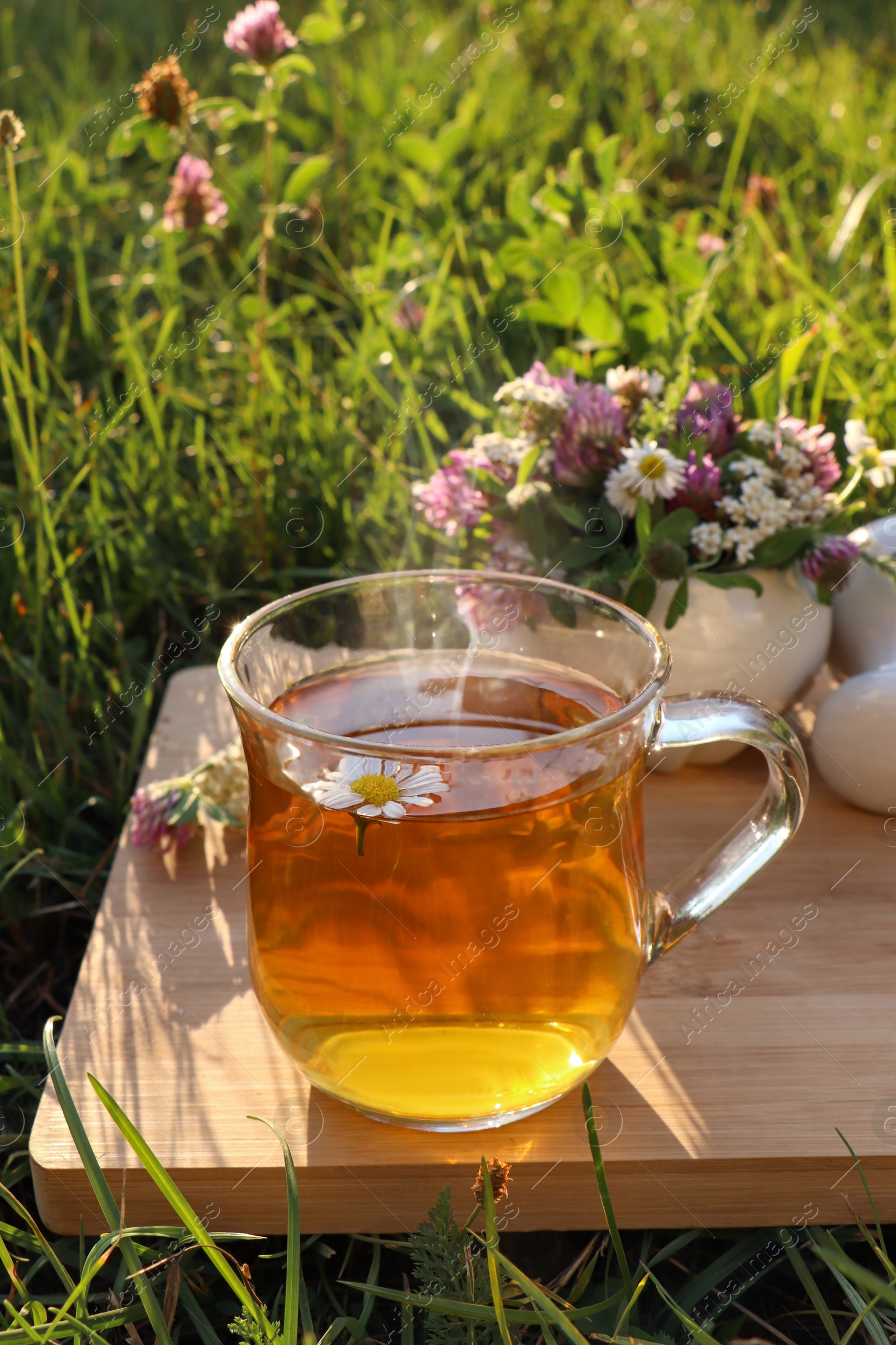 Photo of Cup of aromatic herbal tea, pestle and ceramic mortar with different wildflowers on green grass outdoors