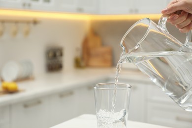 Photo of Woman pouring water from jug into glass at white table in kitchen, closeup. Space for text