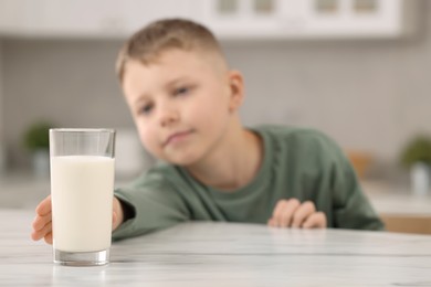Cute boy reaching out for glass of milk at white table in kitchen, selective focus