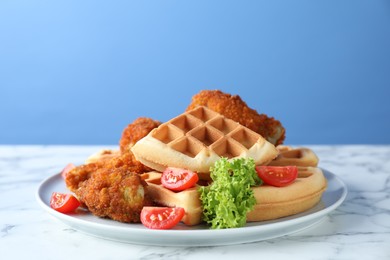 Tasty Belgian waffles served with fried chicken, tomatoes and lettuce on white marble table against light blue background
