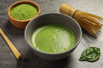 Photo of Cup of fresh matcha tea, green powder, spoon and bamboo whisk on dark grey table, closeup