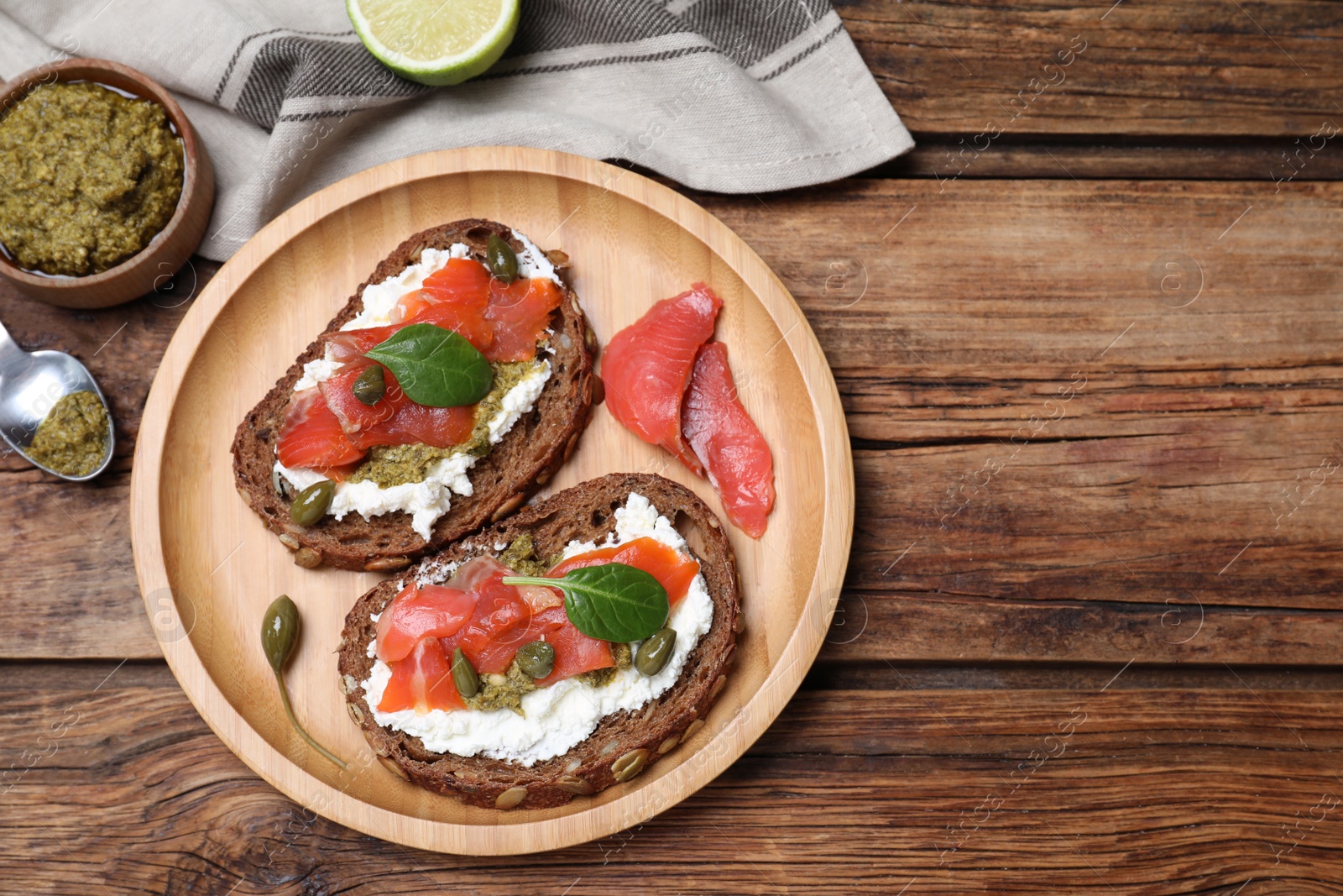 Photo of Delicious sandwiches with cream cheese, salmon, capers and pesto served on wooden table, flat lay. Space for text