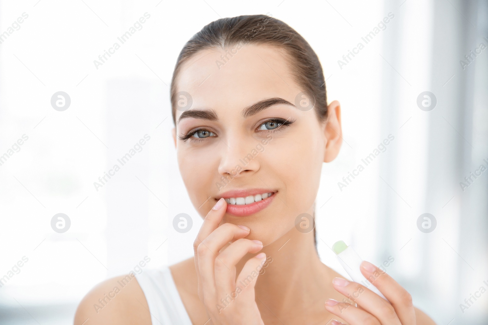 Photo of Young woman applying balm on her lips indoors