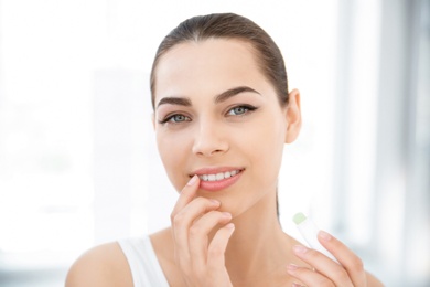 Photo of Young woman applying balm on her lips indoors