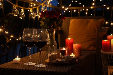 Vase with roses, glasses of wine, snacks and candles on rattan table at balcony in night