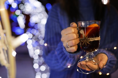 Photo of Woman with glass cup of mulled wine and garland at winter fair, closeup. Space for text