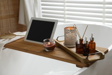Wooden tray with tablet, cosmetic products and burning candles on bath tub in bathroom