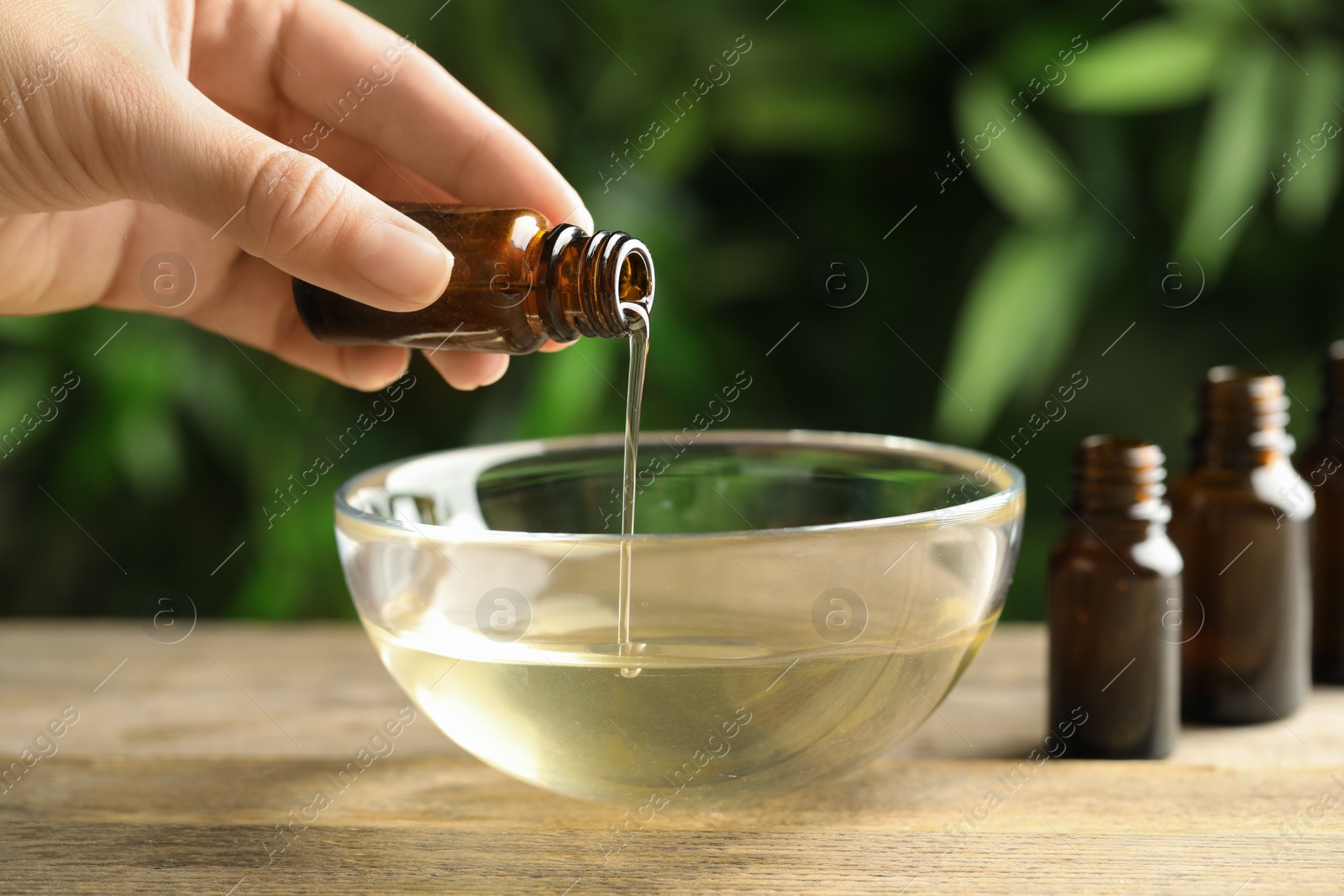 Photo of Woman pouring essential oil from glass bottle into bowl on table, closeup