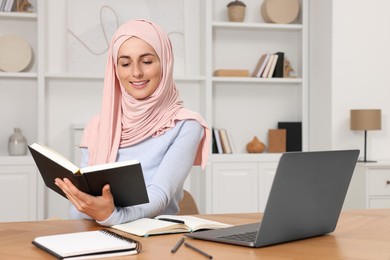Photo of Muslim woman studying near laptop at wooden table in room