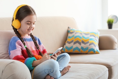 Cute child with headphones and mobile phone on sofa indoors. Space for text