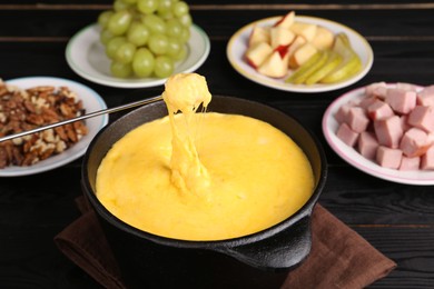 Photo of Dipping piece of ham into fondue pot with melted cheese on black wooden table, closeup