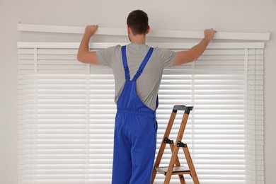 Worker in uniform installing horizontal window blinds on stepladder indoors, back view