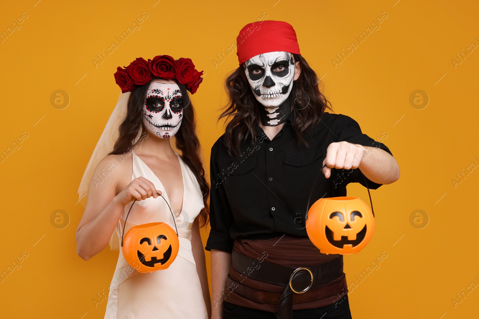 Photo of Couple in scary bride and pirate costumes with pumpkin buckets on orange background. Halloween celebration