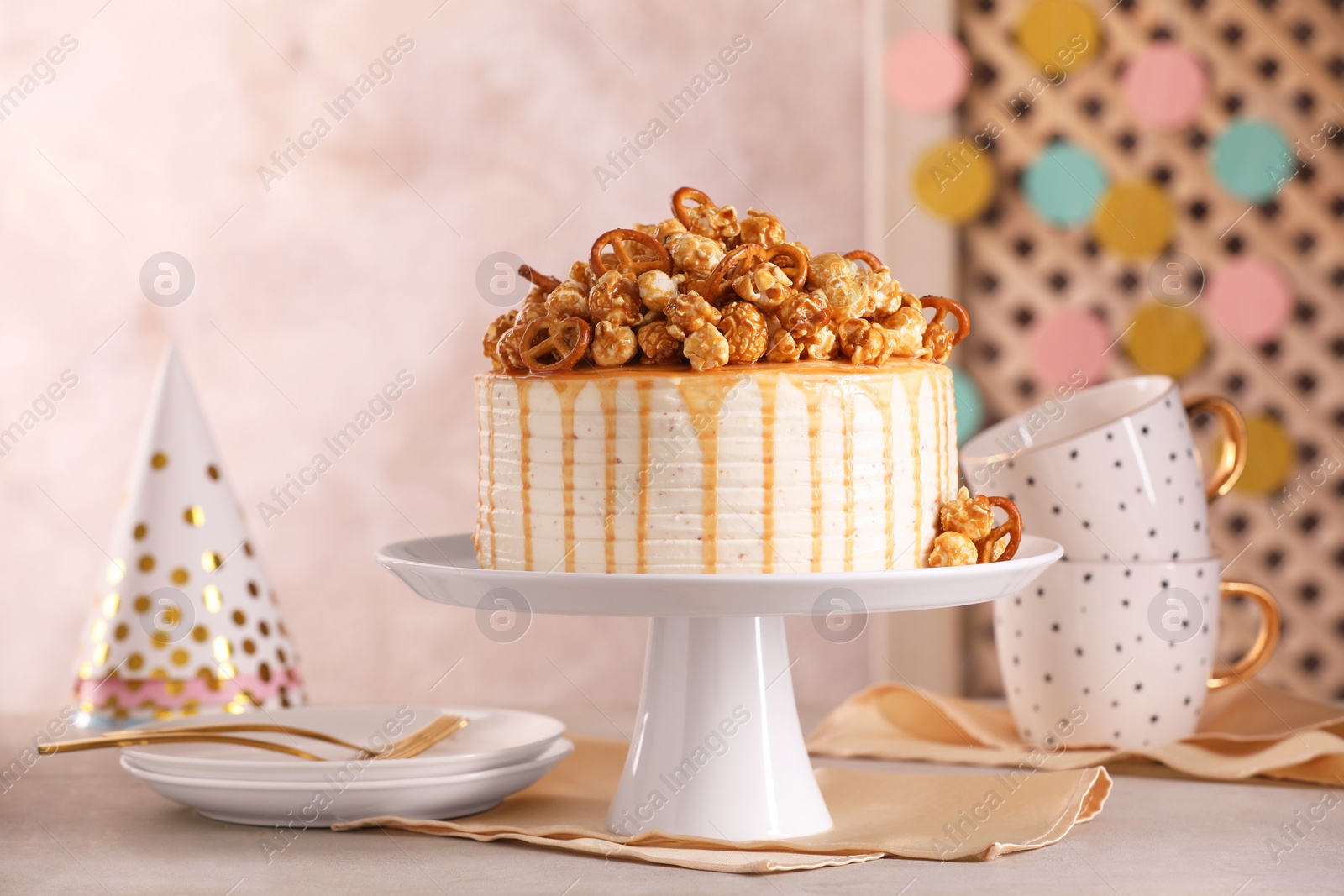 Photo of Caramel drip cake decorated with popcorn and pretzels near tableware on light grey table