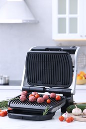 Photo of Electric grill with meatballs, tomatoes and vegetables on white table in kitchen