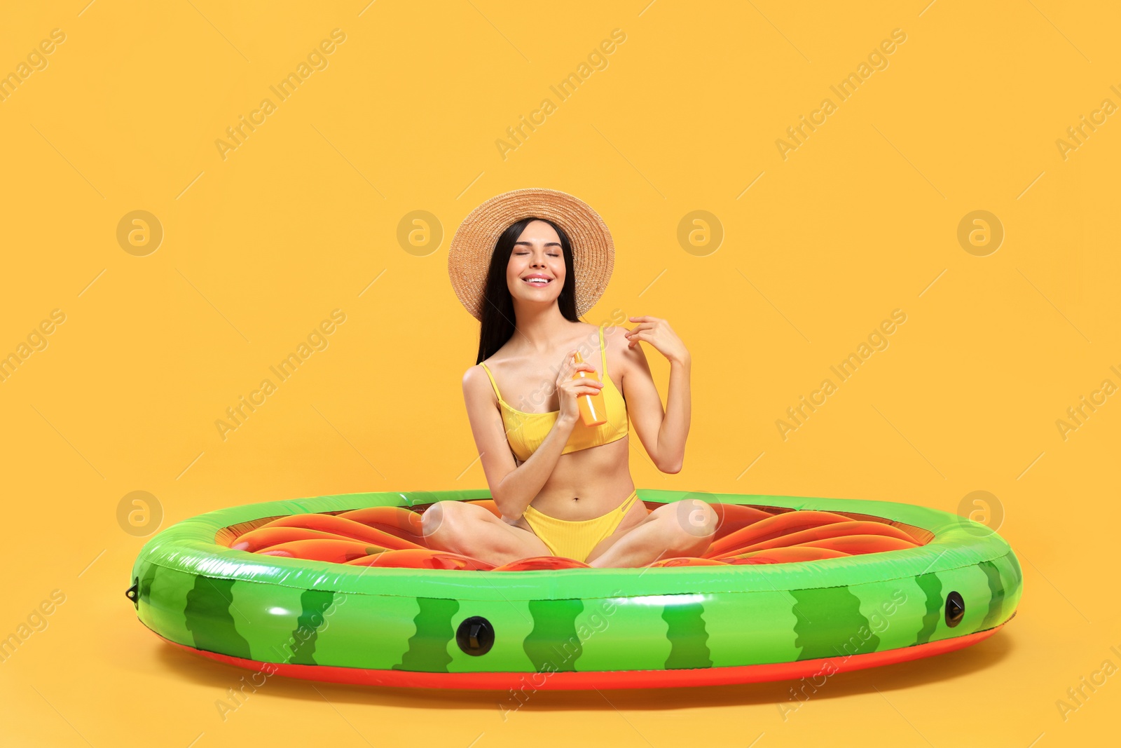 Photo of Happy young woman with sunscreen bottle on inflatable mattress against orange background. Seasonal suntan